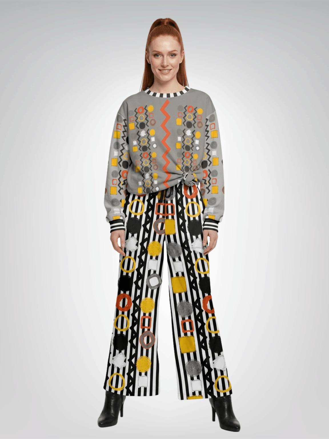 A model stands against a light gray studio background wearing a gray long-sleeve sweatshirt top with colorful geometric patterns and black wide-leg pants with bold black-and-white stripes and circular motifs.