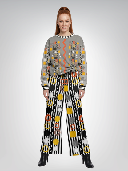A model stands against a light gray studio background wearing a gray long-sleeve sweatshirt top with colorful geometric patterns and black wide-leg pants with bold black-and-white stripes and circular motifs.